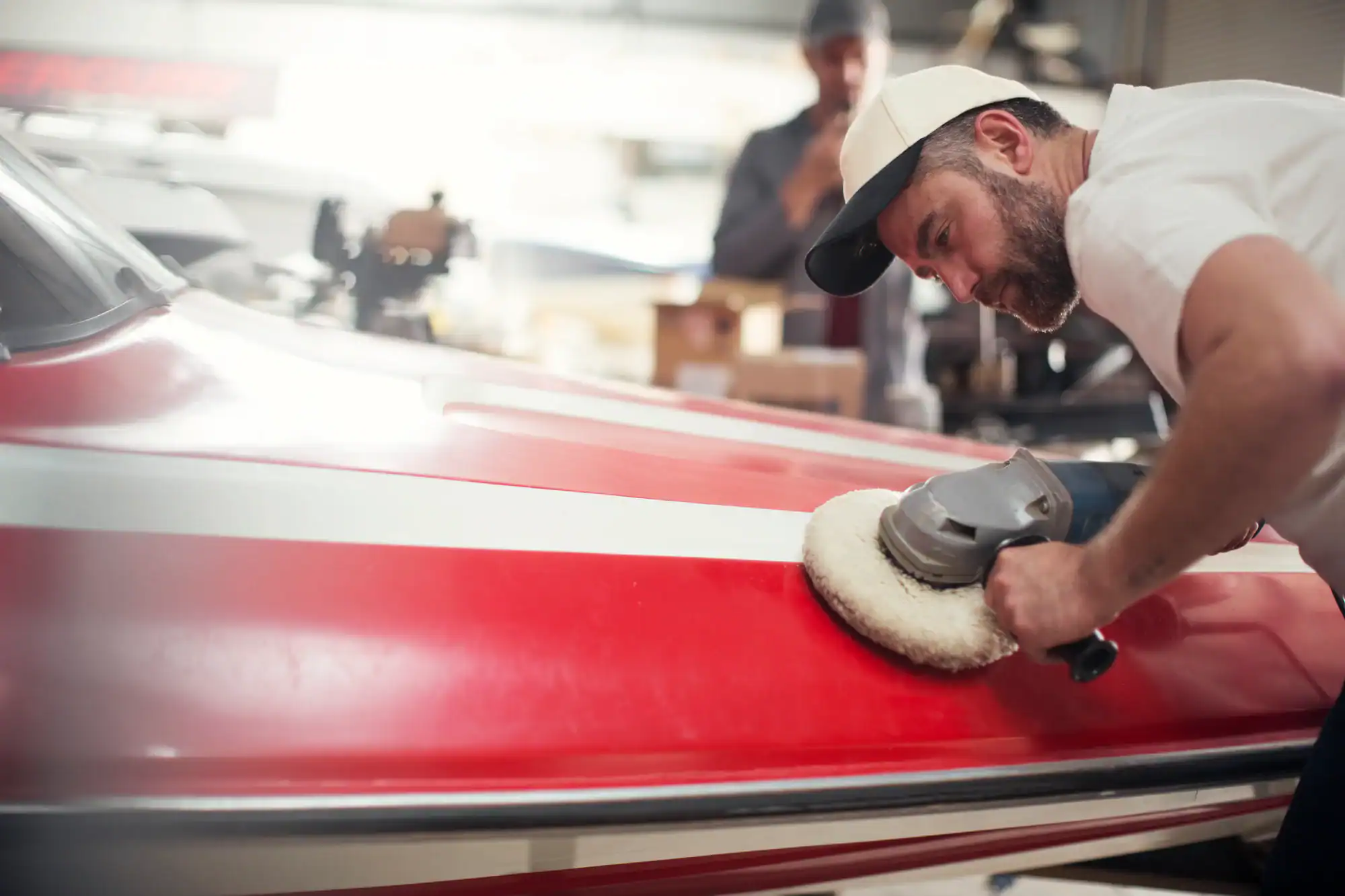 A man in a white cap and t-shirt is polishing the red exterior of a boat with a power buffer at a NY Vehicle Storage Nassau & Suffolk County workshop, while another person is blurred in the background.