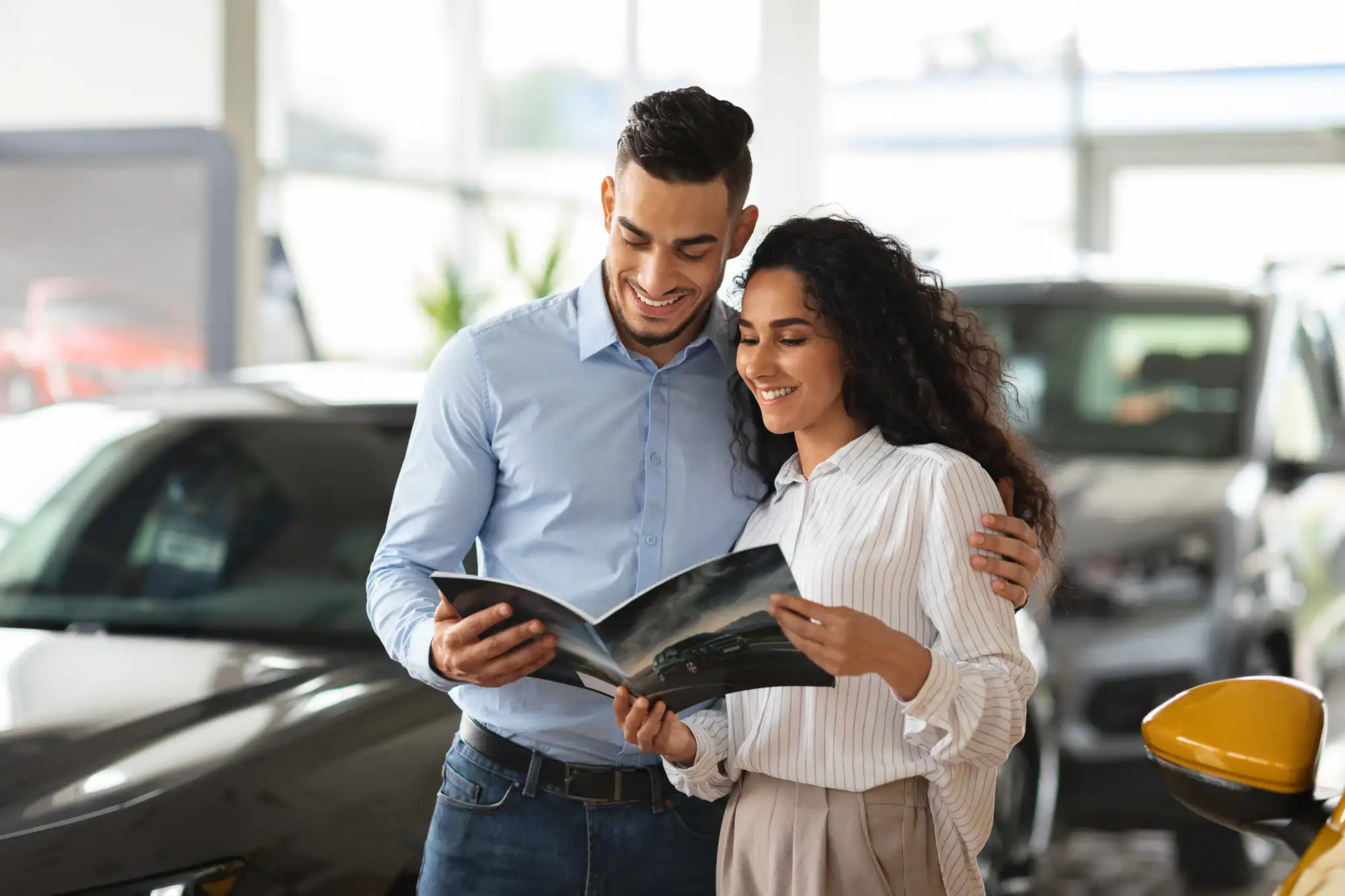 A smiling couple stands together in a car showroom, looking at a brochure. The man has his arm around the woman as they discuss options for Vehicle Storage in Nassau & Suffolk County, NY, with several cars visible in the background.