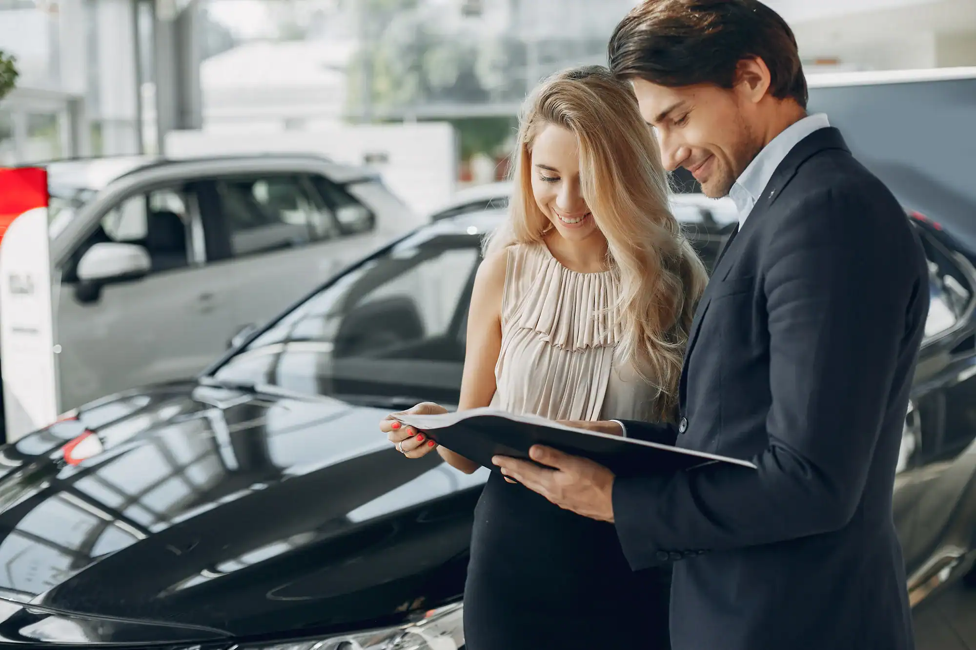 A man in a suit and a woman holding a document stand next to a black car in a NY showroom, smiling as they review Vehicle Storage Nassau & Suffolk County paperwork together.