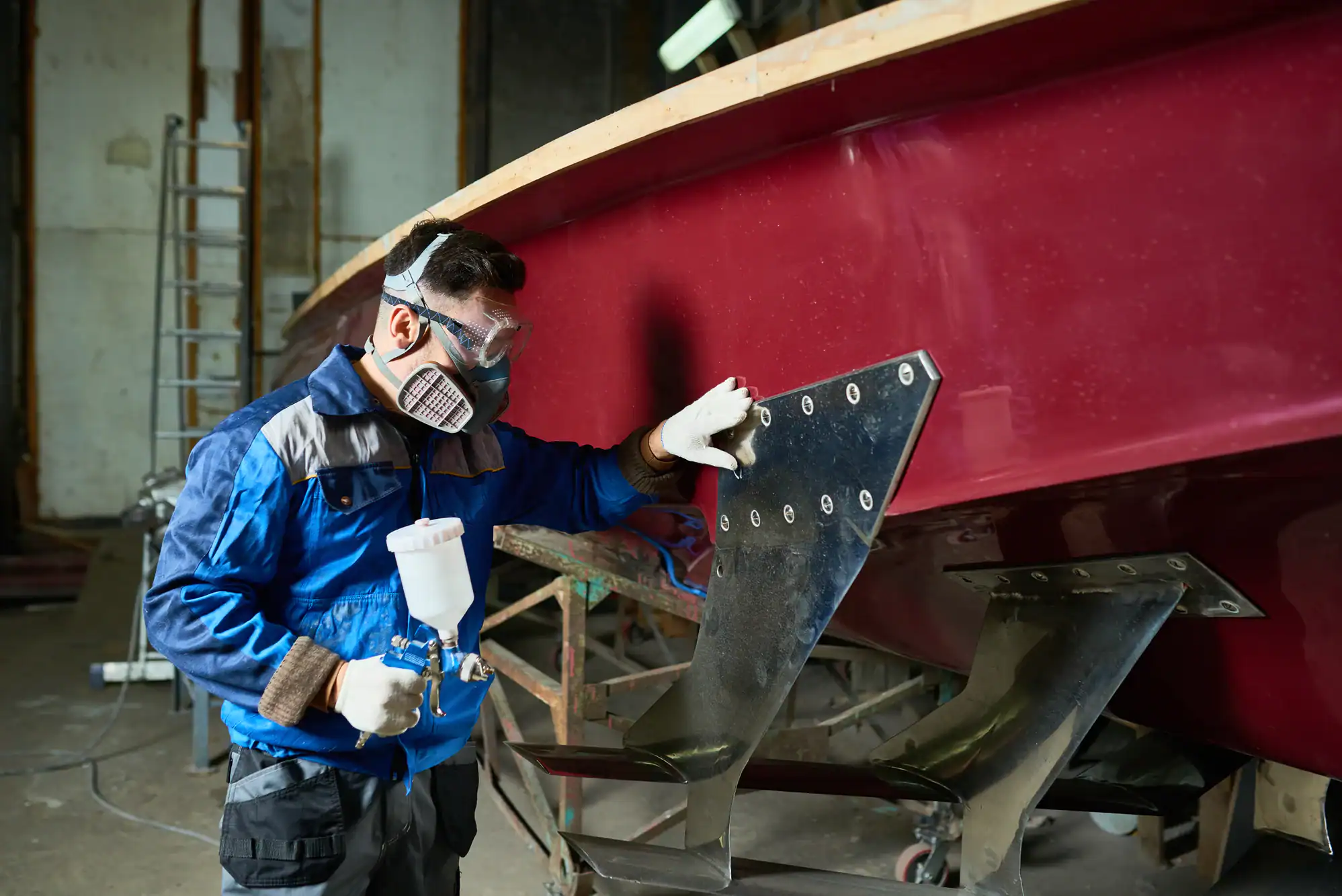In an indoor workshop in NY, a person wearing protective gear and a respirator uses a spray gun to paint or coat the metal parts of a large red boat, often prepared for Vehicle Storage Nassau & Suffolk County.