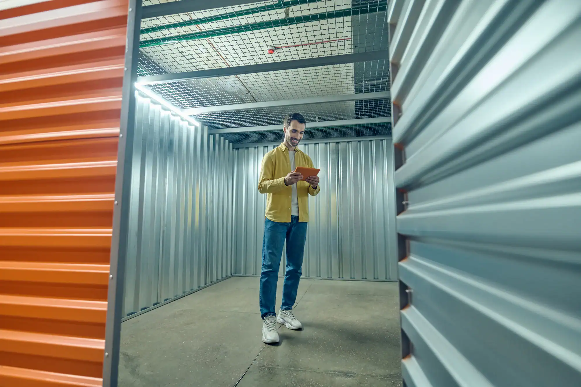 A man stands inside a large, empty storage unit with metal walls, holding and looking at a tablet. He is casually dressed in a yellow jacket, blue jeans, and white sneakers—typical for NY Vehicle Storage Nassau & Suffolk County.