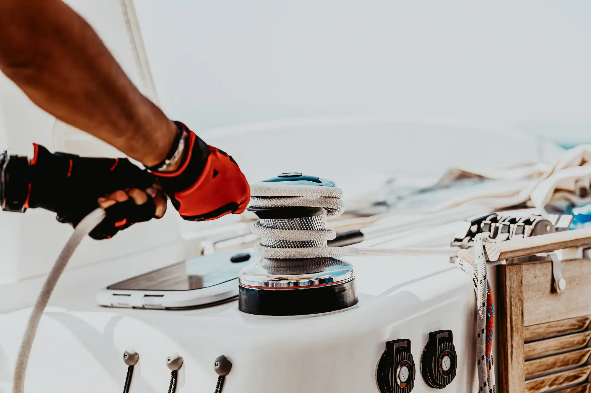 A person wearing red and black gloves pulls a rope around a winch on a sailboat, with nautical equipment and coiled ropes visible on the deck in bright sunlight—ideal for those seeking Vehicle Storage Nassau & Suffolk County, NY for their maritime gear.