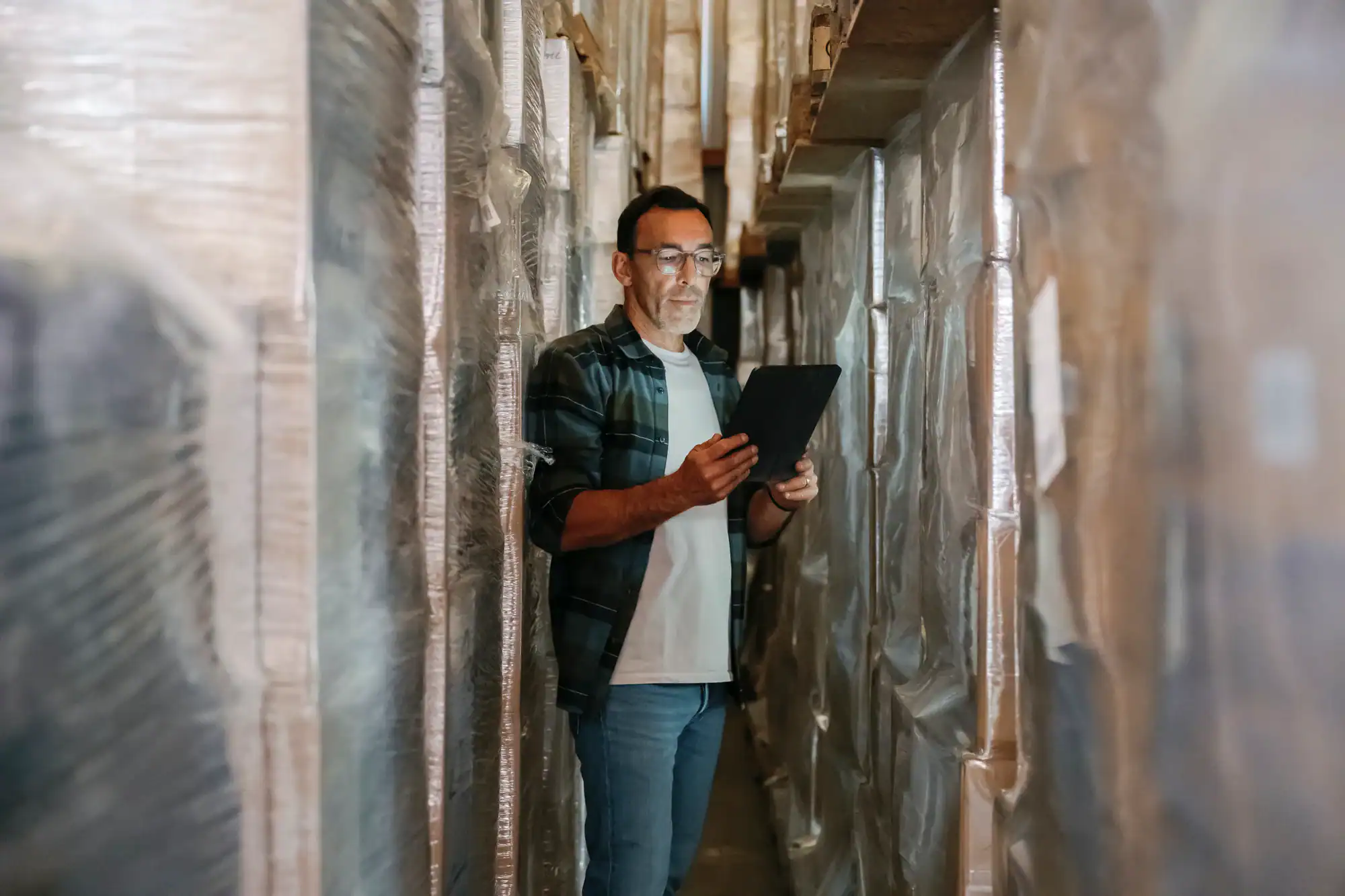 A man stands between tall shelves of wrapped pallets in a NY warehouse, holding and looking at a tablet. He wears glasses, a plaid shirt over a t-shirt, and jeans—perfect for managing Vehicle Storage Nassau & Suffolk County logistics.