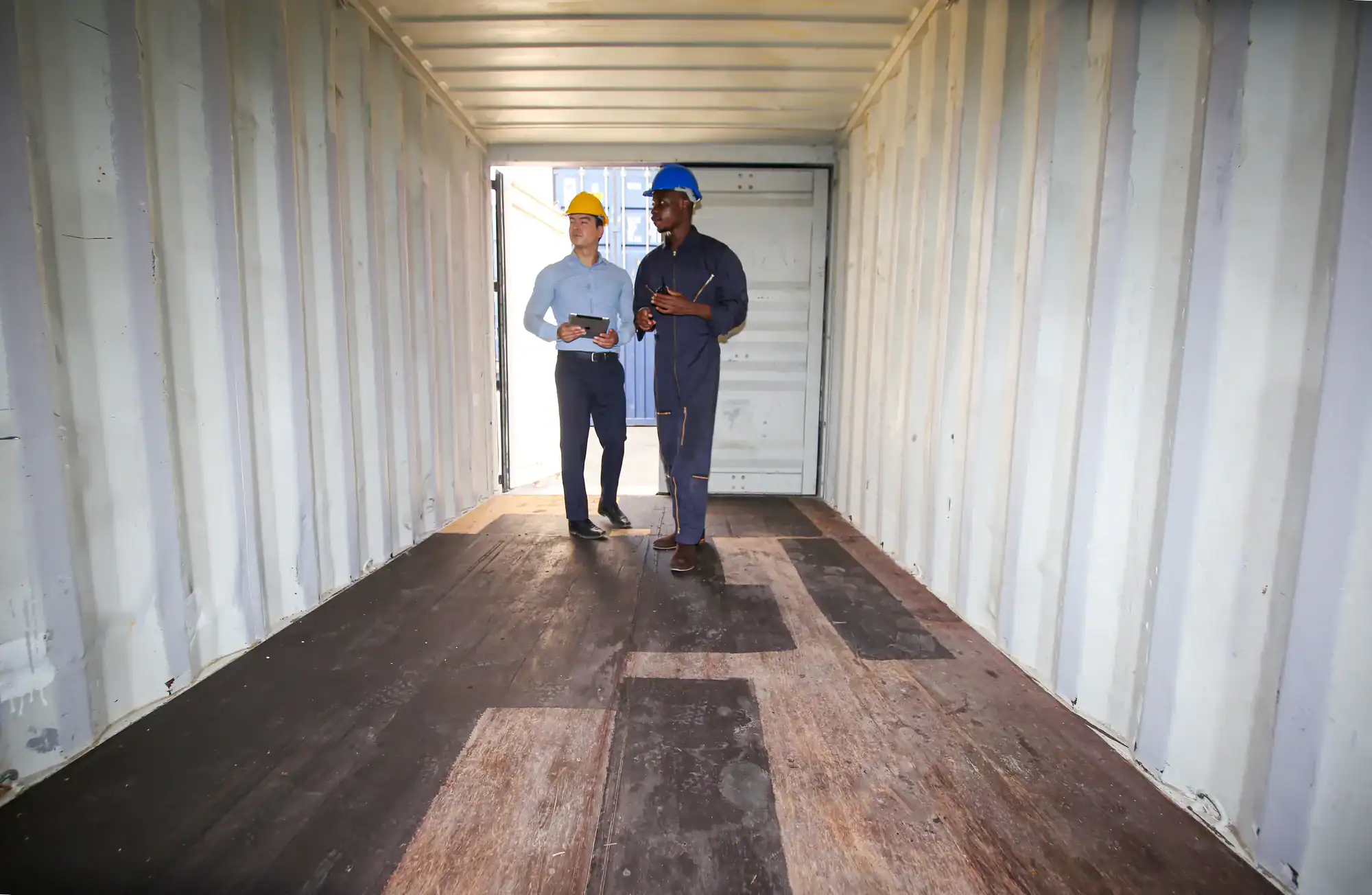 Two men wearing hard hats stand and talk inside an empty shipping container, discussing Vehicle Storage in Nassau & Suffolk County, NY. One is in business attire and the other in work overalls, with sunlight streaming through the open door at the far end.